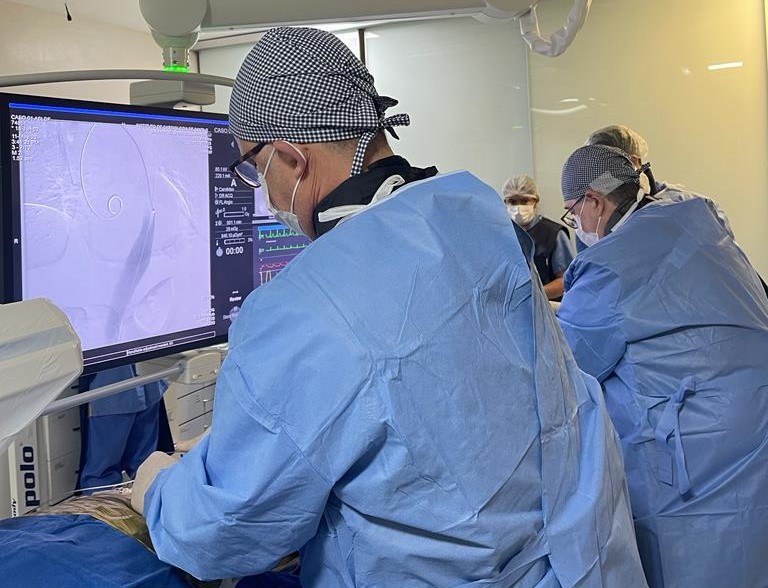 Equipe médica realiza procedimento minimamente invasivo em ambiente cirúrgico. Profissional em primeiro plano opera com auxílio de equipamento de imagem que exibe detalhes anatômicos e sinais vitais do paciente. Todos usam roupas estéreis, toucas e máscaras.