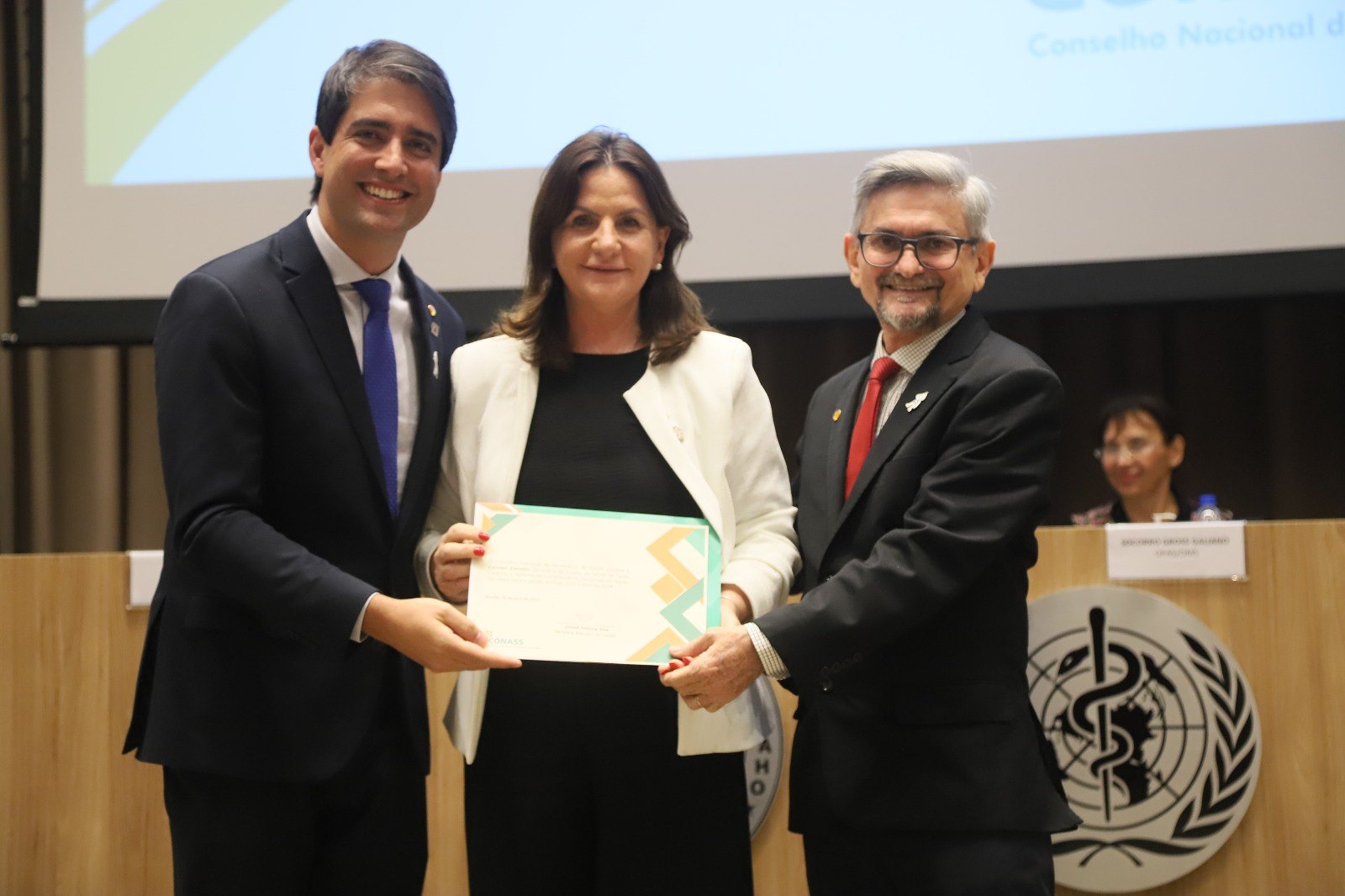 Três pessoas em trajes formais posam sorrindo para uma foto enquanto seguram um certificado. A mulher ao centro veste blazer branco, ladeada por dois homens de terno escuro. Ao fundo, há uma mesa com o logotipo da Organização Mundial da Saúde (OMS) e uma mulher sentada.