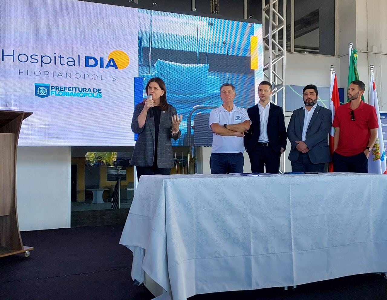 Imagem_do_WhatsApp_de_2023-05-16_às_16.36.58.jpg" alt="Evento da Prefeitura de Florianópolis para o Hospital Dia. Mulher fala ao microfone enquanto cinco homens estão ao fundo, ao lado de bandeiras e de uma tela com logotipo do hospital.