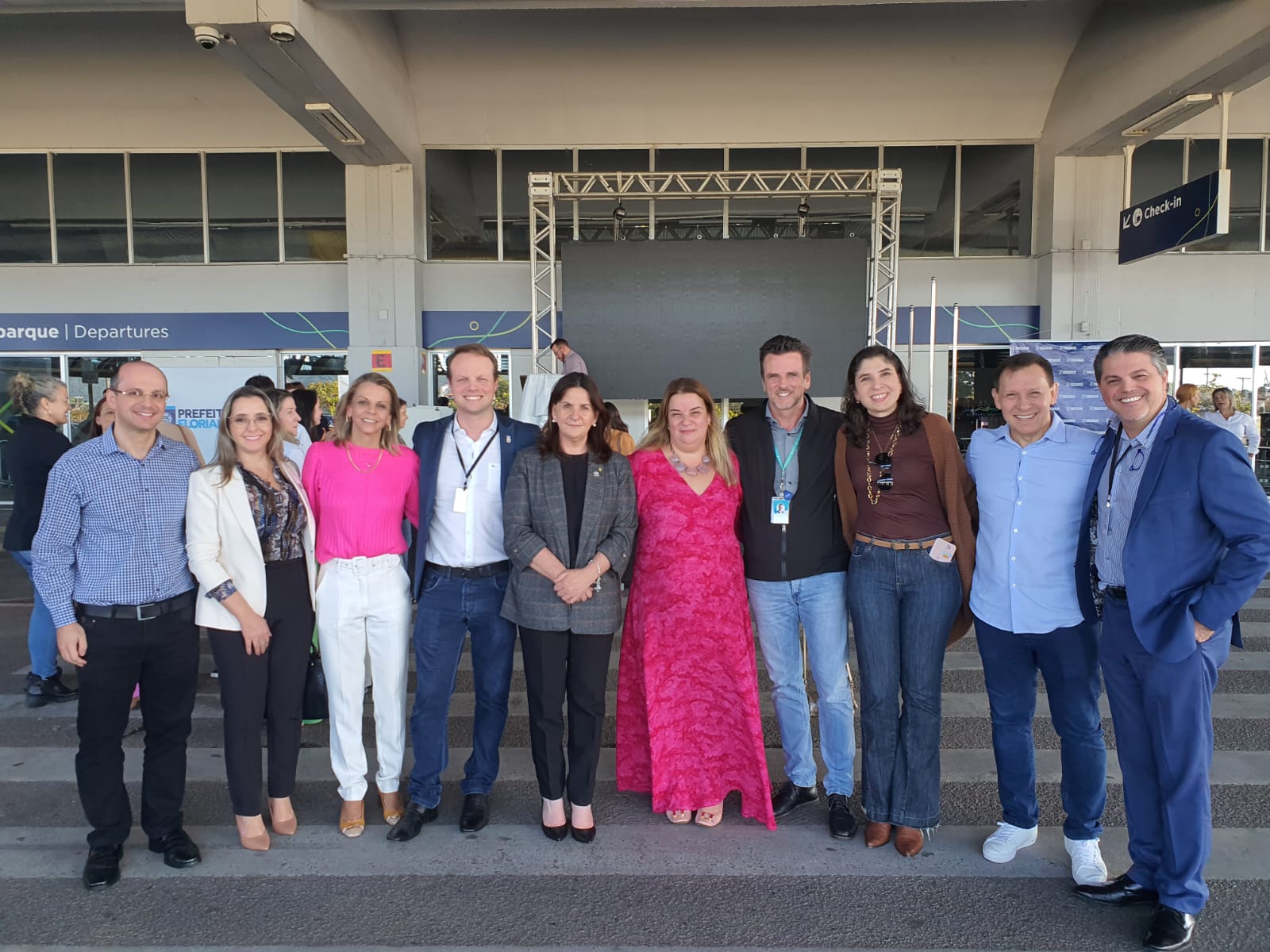 Grupo de pessoas sorrindo em fila, posando para foto em evento oficial da Prefeitura de Florianópolis. Ao fundo, estrutura de palco e sinalização de check-in e embarque, indicando que o evento ocorre em um aeroporto.