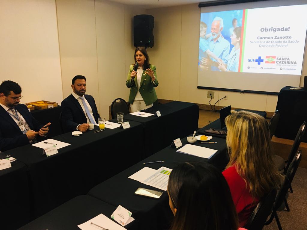 Carmen Zanotto, Secretária de Estado da Saúde de Santa Catarina, fala ao microfone em uma reunião com representantes sentados ao redor de uma mesa em formato de U. Ao fundo, projeção com a mensagem 'Obrigada!' e logotipos do SUS e Governo de Santa Catarina.