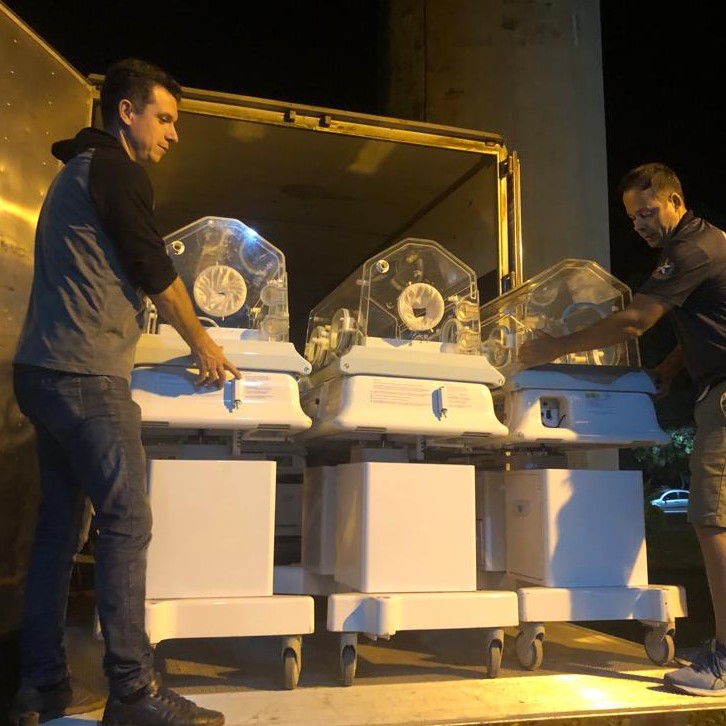 Dois homens descarregam incubadoras neonatais de um caminhão durante a noite. As incubadoras são brancas com estruturas transparentes e estão alinhadas em fileira, prontas para serem transportadas. A cena é iluminada por luzes artificiais.