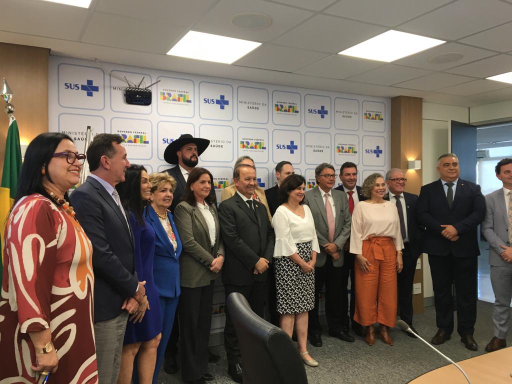 Grupo de autoridades e profissionais da saúde posam para foto oficial em frente a painel com logomarcas do SUS, do Ministério da Saúde e do Governo Federal do Brasil. O cenário é uma sala de reuniões com bandeira do Brasil visível à esquerda. Todos estão sorrindo, em clima de formalidade e colaboração.