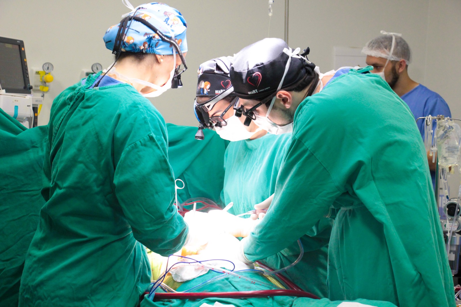 Equipe de cirurgiões vestidos com aventais verdes e toucas cirúrgicas realiza um procedimento em centro cirúrgico. Eles estão concentrados, utilizando equipamentos e luz intensa sobre o campo operatório. Ao fundo, um profissional de saúde observa.