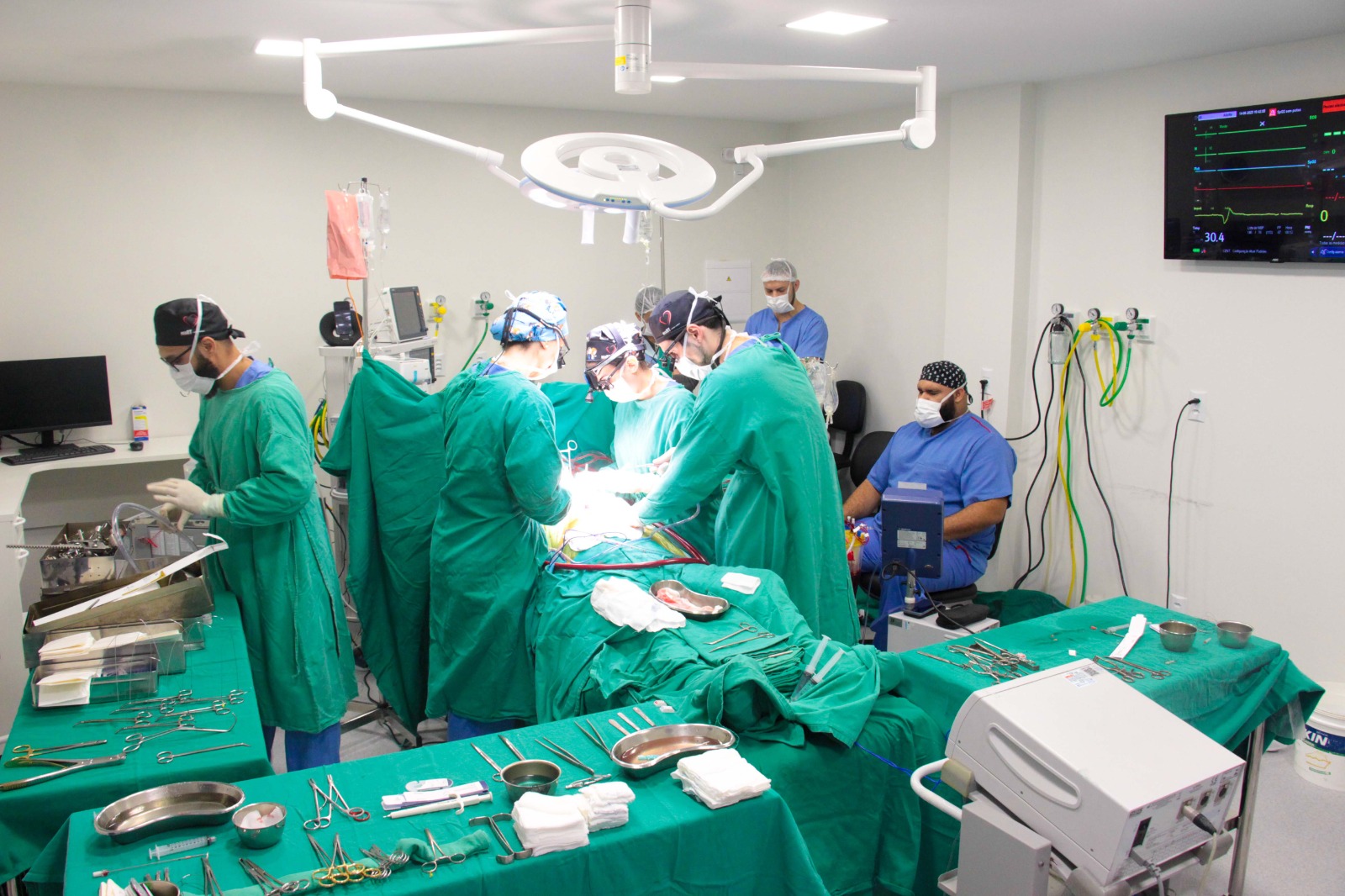 Equipe médica realiza procedimento cirúrgico em centro cirúrgico moderno, com instrumental cirúrgico organizado em mesas esterilizadas e monitor de sinais vitais ligado na parede.