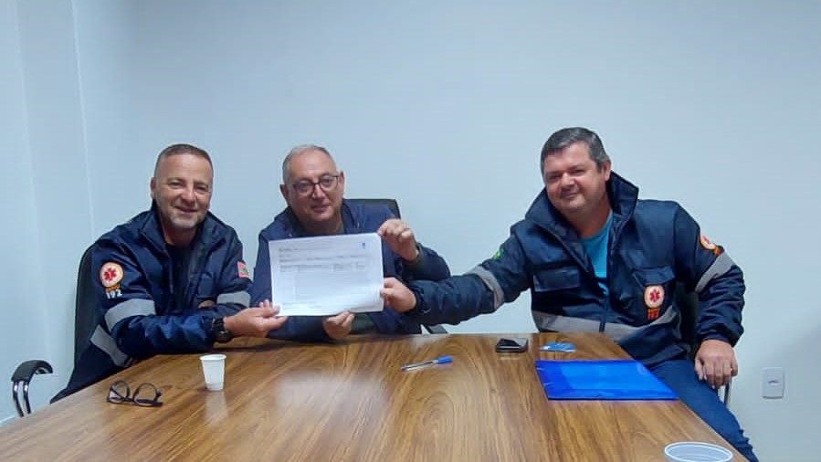 Três homens uniformizados com jaquetas do SAMU, com logotipo e número 192, estão sentados em volta de uma mesa de madeira. Eles sorriem e seguram juntos um documento. Sobre a mesa há copos descartáveis, uma caneta, óculos, celular e uma pasta azul. O ambiente tem paredes brancas e aparência de sala administrativa.