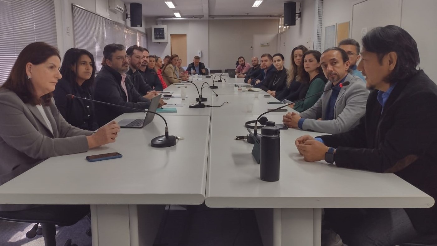 Imagem de uma reunião formal com aproximadamente 20 pessoas, divididas em dois lados de uma mesa retangular comprida. Todas estão sentadas e algumas utilizam microfones fixos na mesa. A maioria está atenta, ouvindo ou conversando. A sala tem janelas com persianas, paredes claras e iluminação fluorescente. Um celular, garrafas térmicas, laptops e cadernos estão sobre a mesa.
