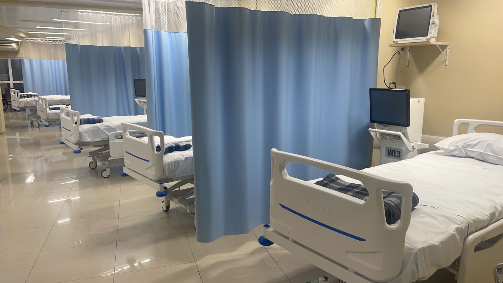 Leito hospitalar moderno em ala recém-reformada, com cortinas azuis dividindo os espaços entre camas equipadas com monitores e colchões. Ambiente limpo, iluminado e organizado.