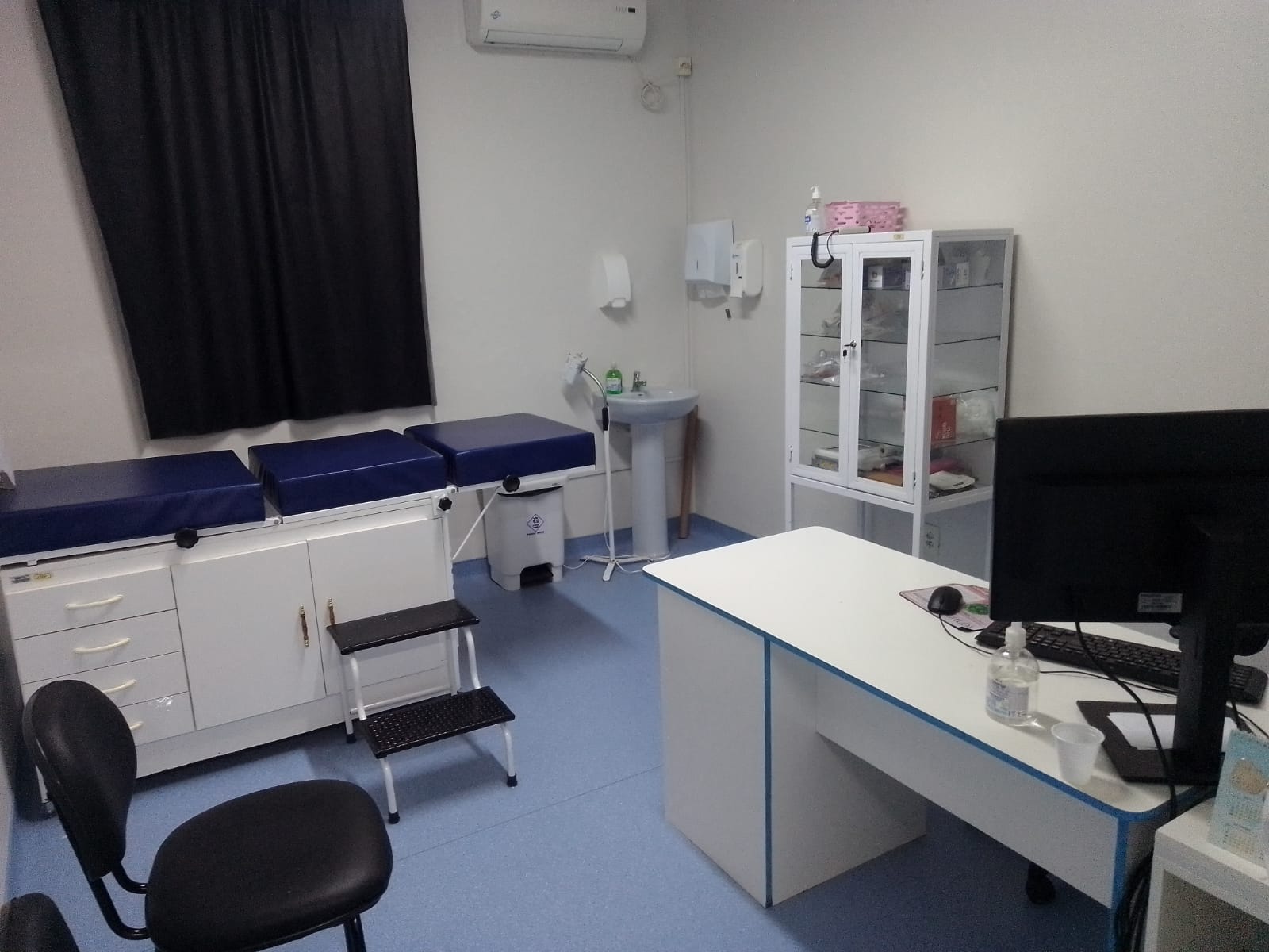 Consultório médico com maca de exames, mesa com computador, cadeiras, armário de vidro com suprimentos e pia de higienização em ambiente limpo e organizado.