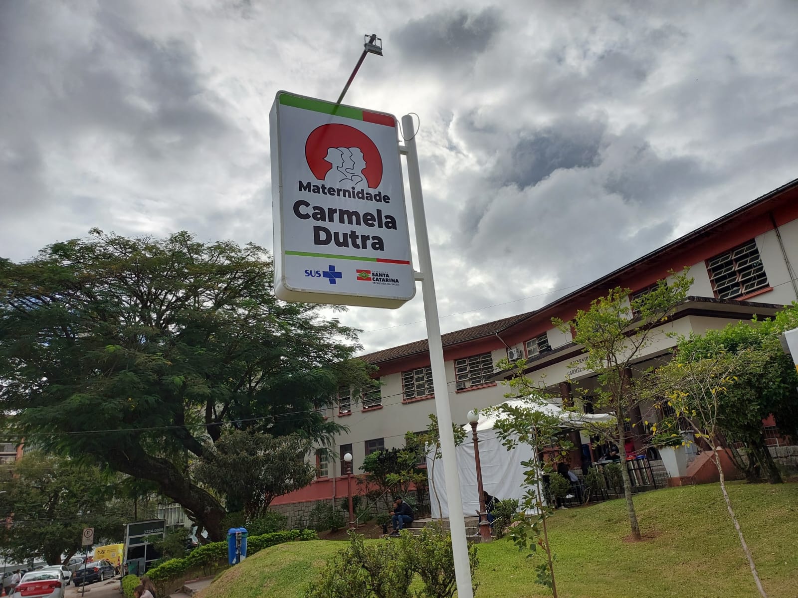Fachada da Maternidade Carmela Dutra, em Florianópolis (SC), com uma placa de identificação em destaque no primeiro plano. A placa traz o logotipo da maternidade e os logos do SUS e do Governo de Santa Catarina. Ao fundo, o prédio da maternidade é visível com suas janelas, árvores ao redor e tendas brancas montadas no jardim em frente à entrada principal. O céu está parcialmente nublado.