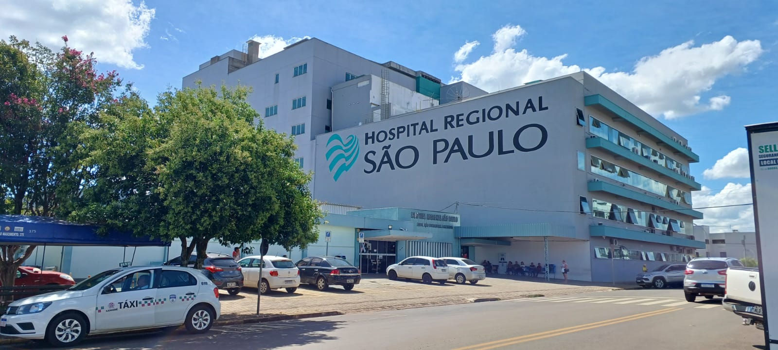 Vista externa do Hospital Regional São Paulo em uma manhã ensolarada. O prédio de vários andares é cinza com detalhes em azul e tem o nome da instituição escrito em letras grandes na fachada. Em frente, há árvores, carros estacionados e um táxi branco parado na rua.