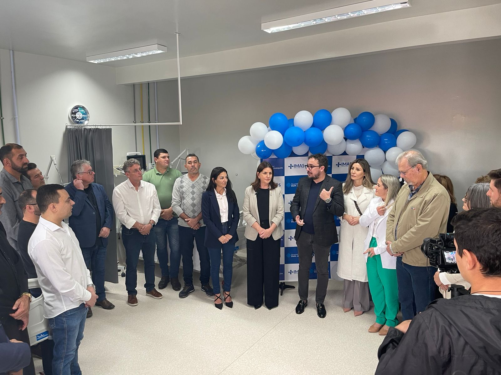 Grupo de autoridades e profissionais de saúde em cerimônia de inauguração no Hospital Dom Joaquim, com balões azuis e brancos ao fundo e painel do IMAS. Um homem ao centro faz um discurso enquanto os demais ouvem atentamente.