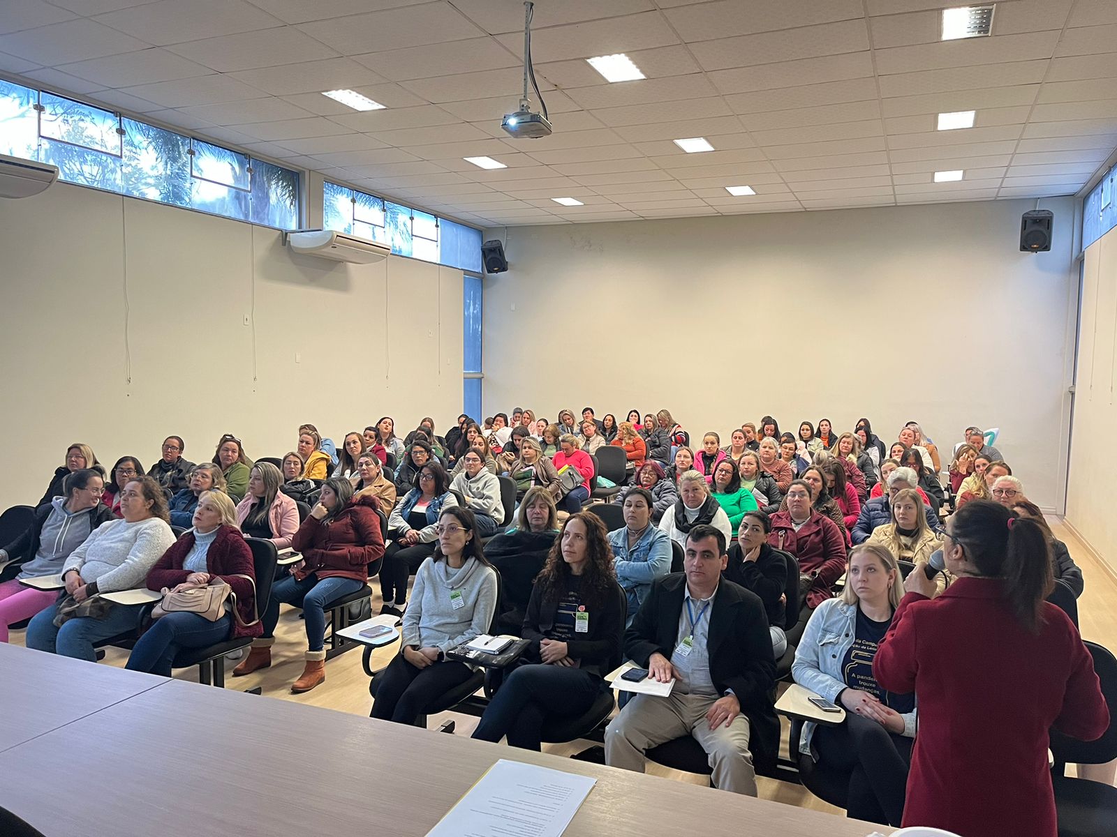 Auditório cheio com dezenas de profissionais da saúde assistindo a palestra; mulher de casaco vermelho fala ao microfone de frente para o público.