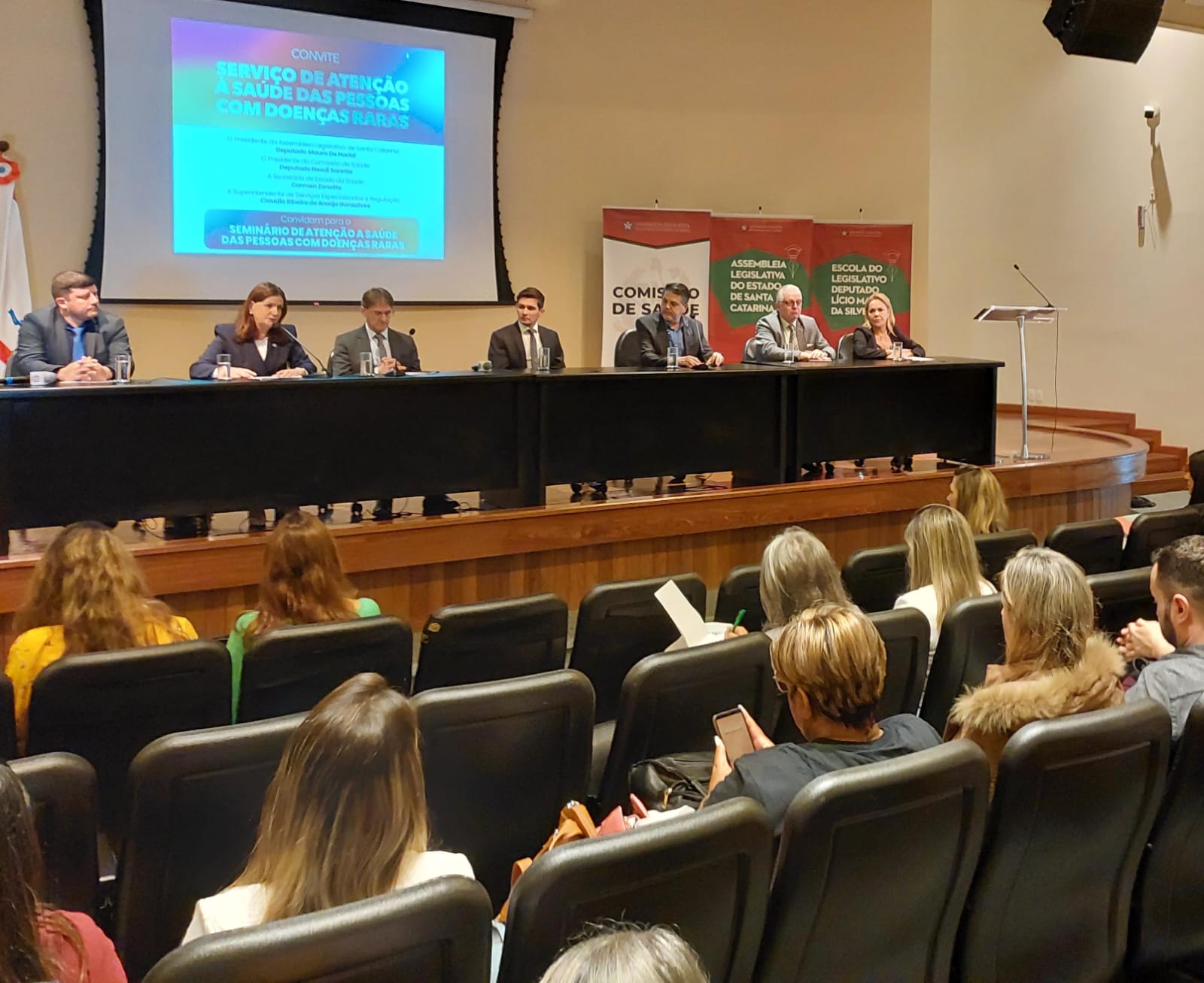 Autoridades sentadas em mesa de seminário sobre atenção à saúde de pessoas com doenças raras, com telão ao fundo e público assistindo no auditório da Assembleia Legislativa de Santa Catarina.
