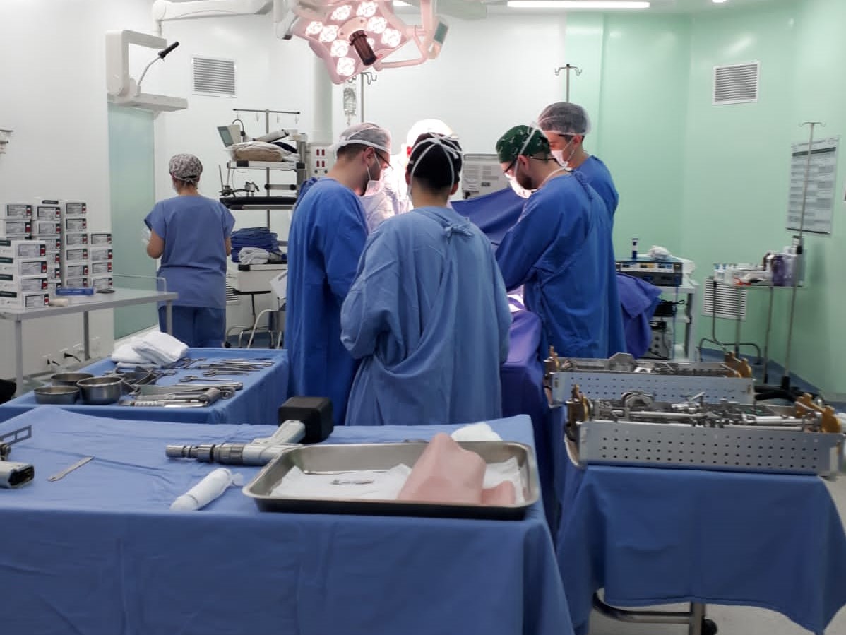Equipe médica com aventais e toucas cirúrgicas realiza um procedimento em uma sala de cirurgia equipada. Em primeiro plano, mesas com instrumentos cirúrgicos organizados sobre campos estéreis.
