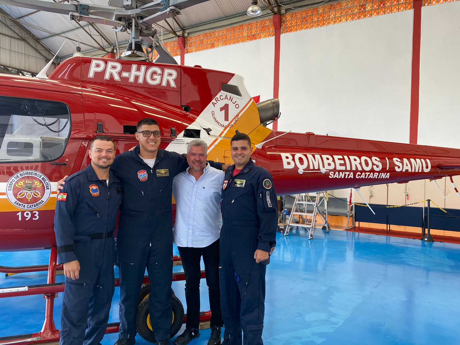 Quatro homens posam sorrindo em frente a um helicóptero vermelho do Corpo de Bombeiros Militar e SAMU de Santa Catarina, com a inscrição "Arcanjo 1" visível. Três usam macacões de voo e um está de camisa branca. Estão em um hangar com piso azul.