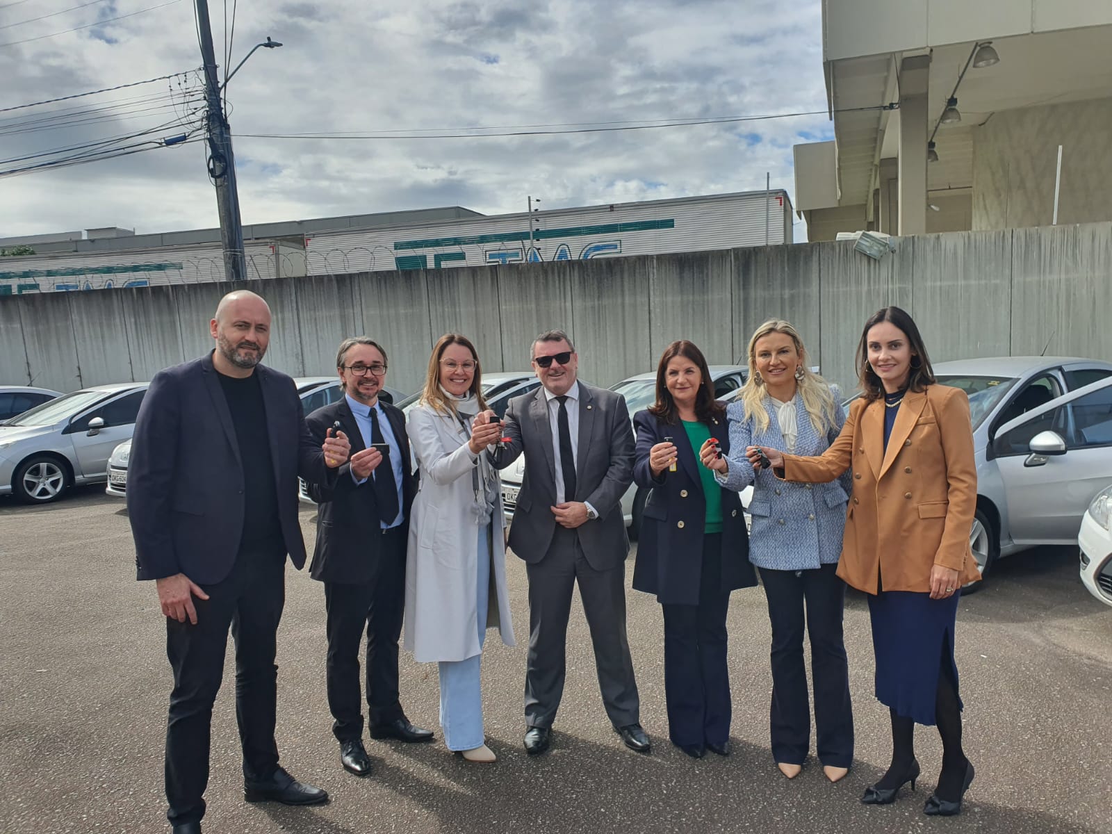 Grupo de sete pessoas, incluindo homens e mulheres trajando roupas sociais, posando em área externa de estacionamento, segurando chaves de veículos. Ao fundo, diversos carros prateados estão estacionados. A cena sugere entrega oficial de veículos para uso institucional ou governamental.