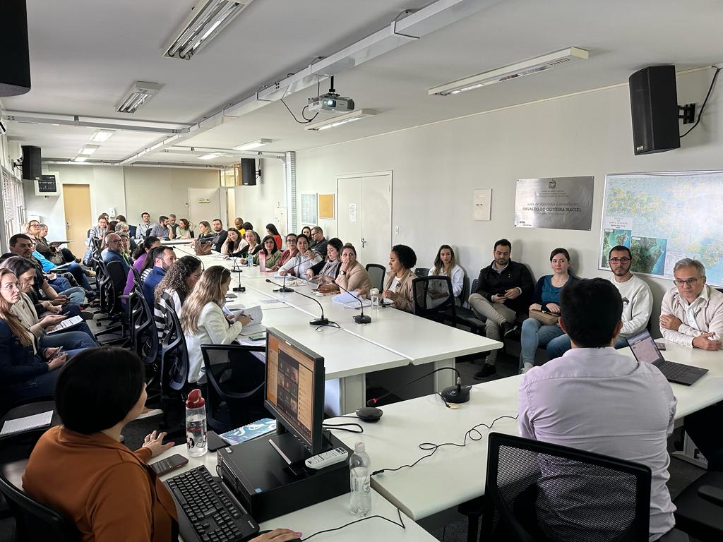 Sala de conferência repleta de pessoas sentadas ao redor de uma grande mesa em formato de U. Algumas estão com papéis e laptops, outras escutam atentas. Há microfones sobre as mesas, um projetor no teto e uma placa na parede indicando o nome da sala. O clima é de reunião ou capacitação técnica.