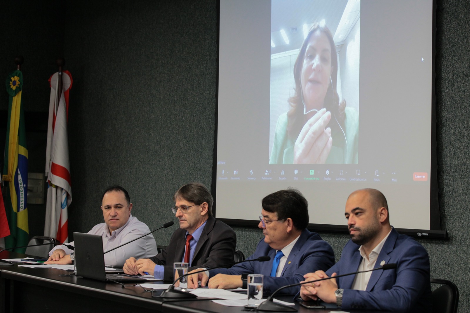Mesa composta por quatro homens em trajes formais durante audiência pública; ao fundo, projeção de uma mulher participando por videoconferência. Bandeiras do Brasil e de estados ao lado.