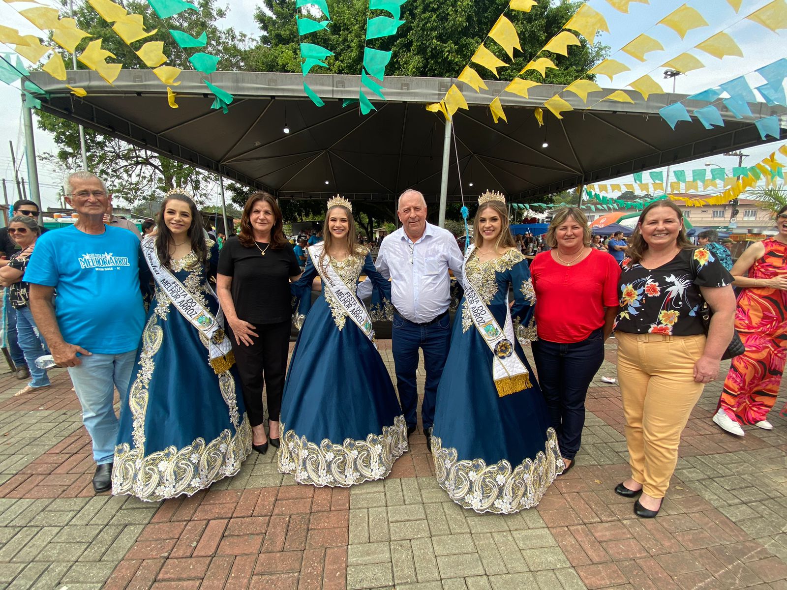 Rainhas vestidas com trajes típicos azul e dourado posam ao lado de autoridades e visitantes sob decoração com bandeirinhas coloridas em evento ao ar livre, realizado em praça pública com barracas ao fundo.
