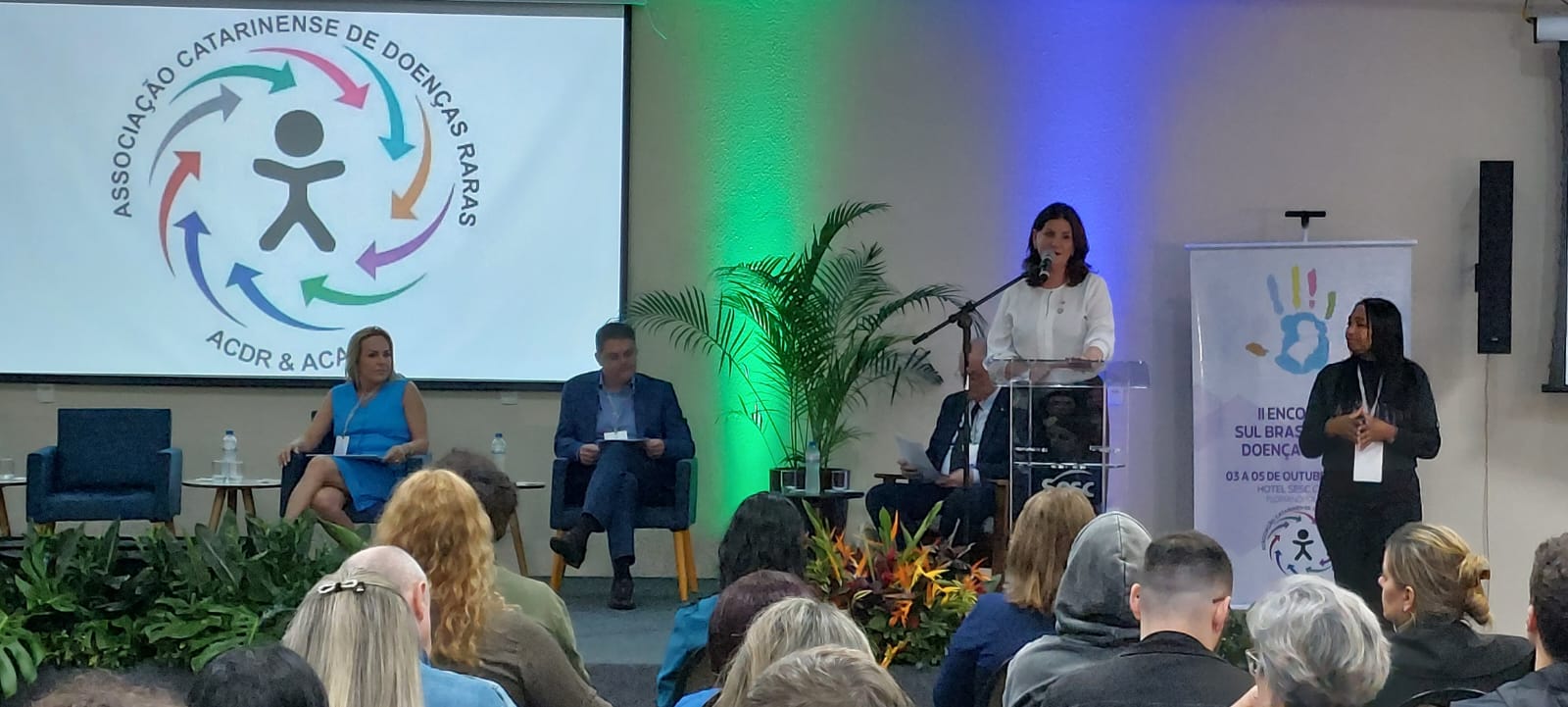 Auditório com público assistindo a uma apresentação. No palco, uma mulher em pé fala ao microfone em púlpito transparente, enquanto outra ao lado faz tradução em Libras. Ao fundo, há um telão com o logotipo da Associação Catarinense de Doenças Raras e um banner com o nome do evento.