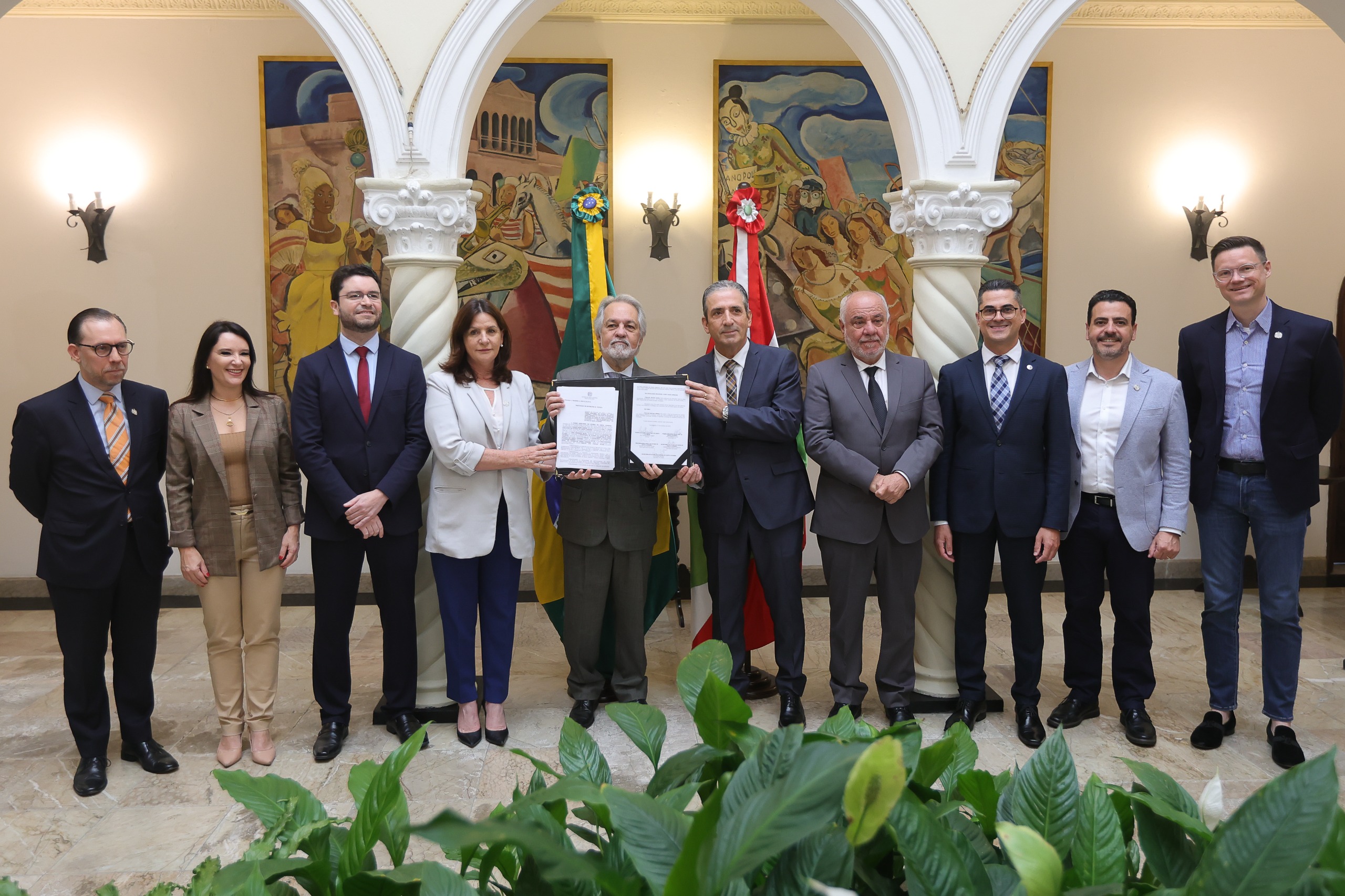 Grupo de nove pessoas posando para foto em ambiente formal, com dois homens ao centro segurando documento aberto, bandeiras do Brasil e de estado ao fundo e murais coloridos nas paredes.