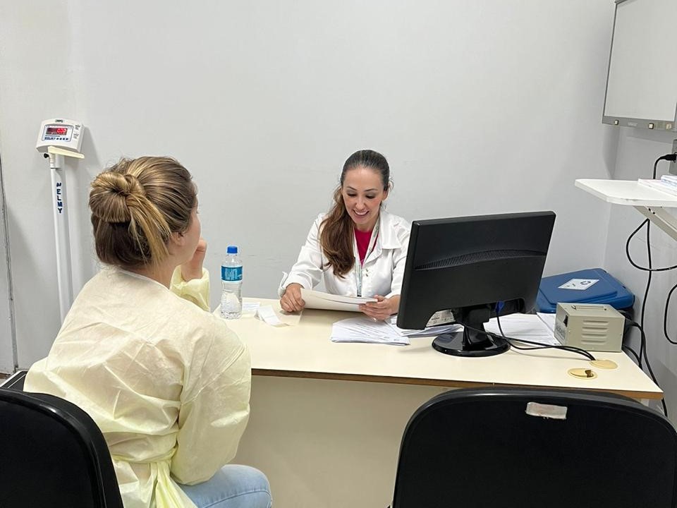 Profissional de saúde, usando jaleco branco, atende paciente em consultório, analisando documentos e sorrindo, com computador e materiais sobre a mesa.