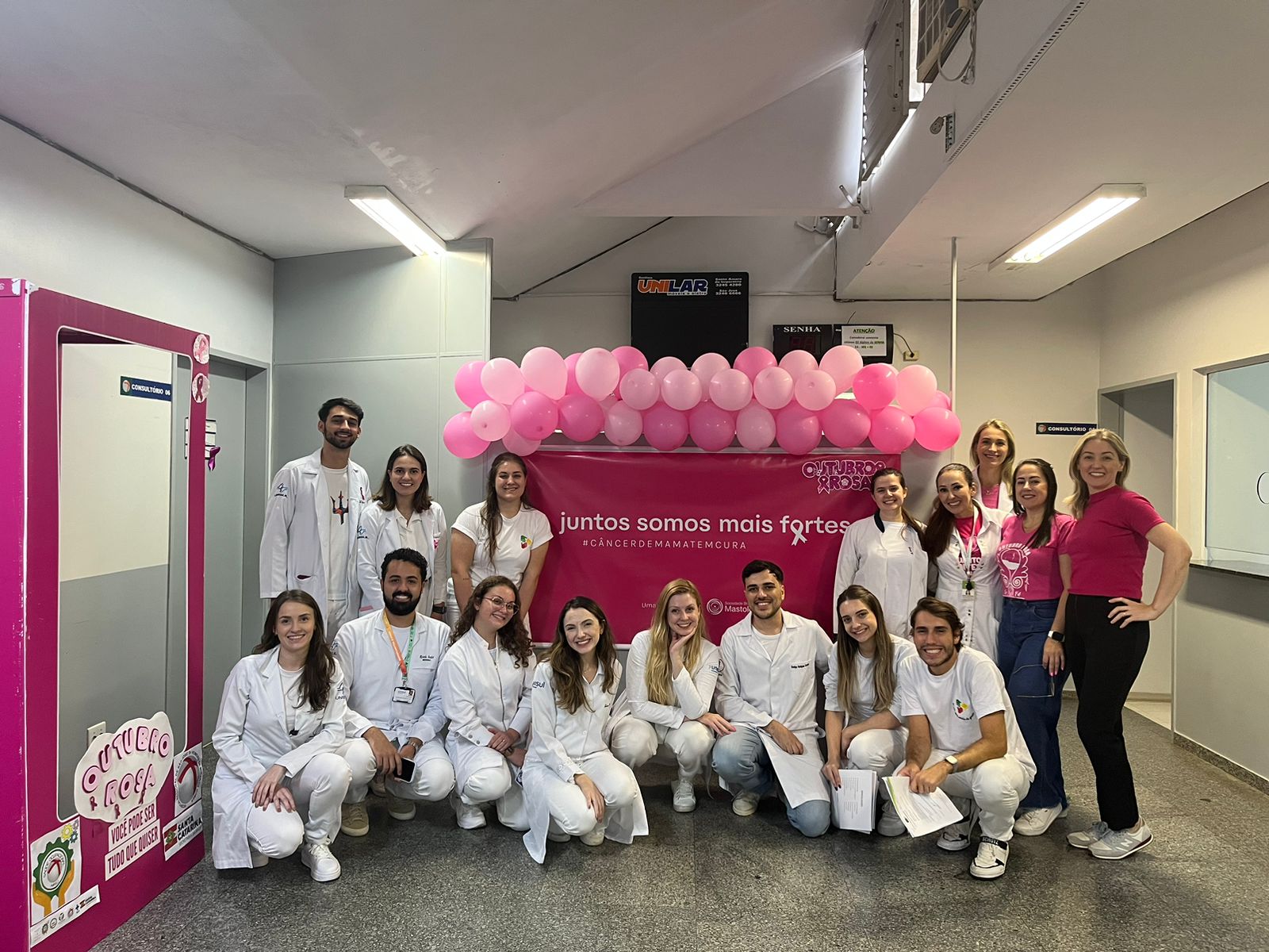 Grupo de profissionais de saúde e voluntários, vestidos de branco e rosa, posando em frente a um painel decorado com balões cor-de-rosa e a frase “Juntos somos mais fortes – #CâncerDeMamaTemCura”, em ação de conscientização do Outubro Rosa.