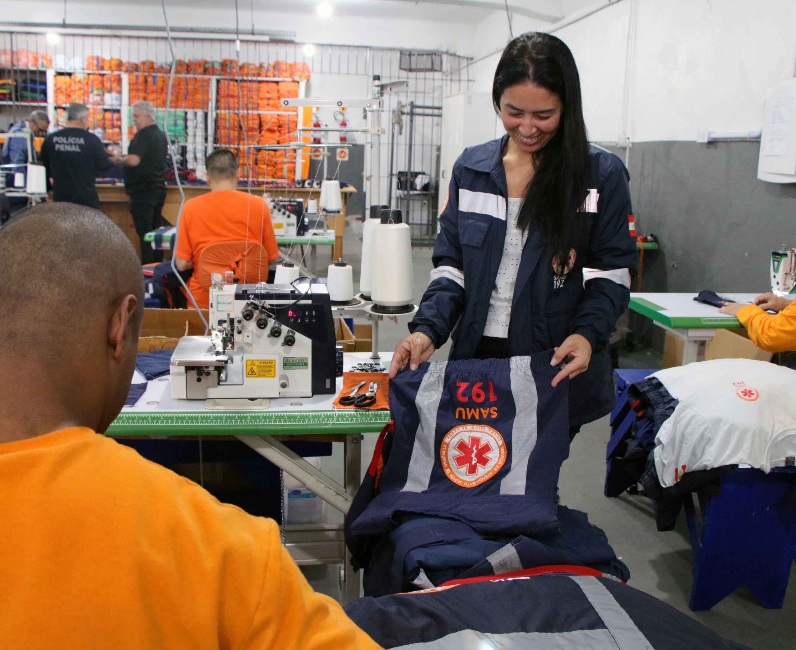 Mulher segurando uniforme azul do SAMU 192 em oficina de costura, com detentos trabalhando em máquinas de costura ao fundo.