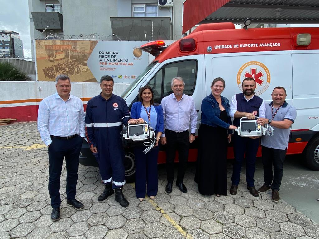 Grupo de sete pessoas posando em frente a uma ambulância do SAMU, com dois profissionais segurando monitores/desfibriladores recém-entregues.