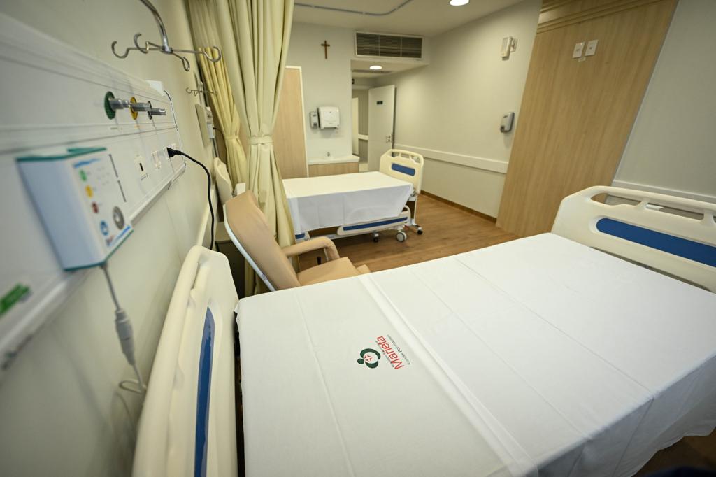 A imagem mostra um quarto hospitalar moderno e bem iluminado. Há duas camas com lençóis brancos estampados com o logotipo do Hospital Marieta, cada uma equipada com cabeceira hospitalar e pontos de oxigênio, ar comprimido e tomadas para equipamentos médicos. Entre as camas, há uma poltrona reclinável para acompanhantes, e ao fundo vê-se outra cama próxima à parede.  O ambiente é organizado, com piso de madeira, cortina divisória para privacidade, dispenser de álcool em gel na parede e um crucifixo acima da porta, transmitindo um ar acolhedor e humanizado.