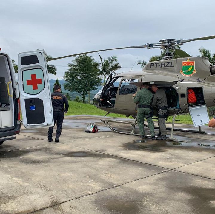 Cena em área aberta mostrando um helicóptero militar de resgate, com a inscrição "PT-HZL" e brasão na fuselagem, estacionado próximo a uma ambulância com a porta traseira aberta e cruz vermelha visível. Dois militares em uniforme verde interagem junto à porta lateral do helicóptero, enquanto uma pessoa de uniforme escuro da equipe de emergência está próxima à ambulância. O cenário é gramado com árvores ao fundo e céu nublado.