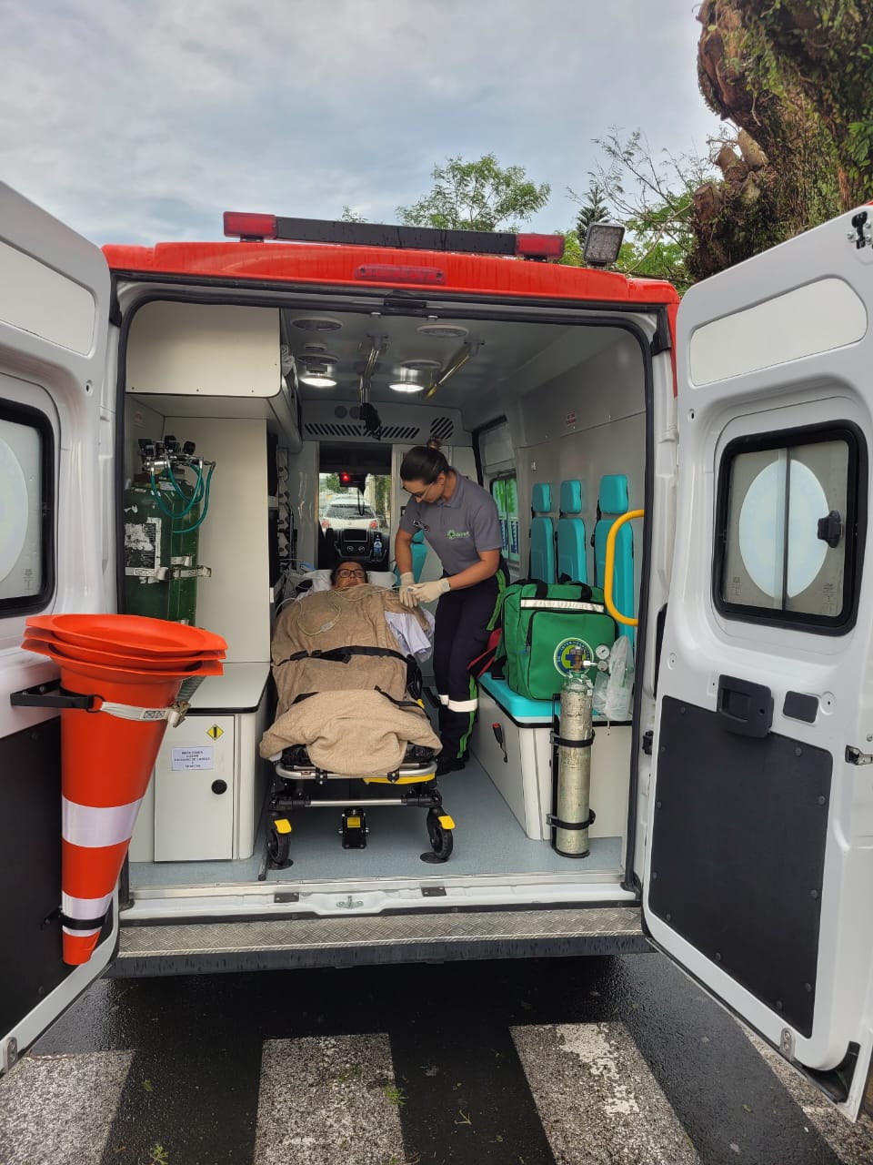 Parte traseira aberta de uma ambulância, revelando o interior equipado com cadeiras, cilindro de oxigênio e armários. Uma paciente está deitada em uma maca coberta por um cobertor bege, sendo atendida por uma profissional de saúde com uniforme cinza e calça com faixas refletivas. Na porta da ambulância, há um cone de sinalização laranja com faixas brancas preso por cinta. Ao fundo, é possível ver parte da rua e árvores.