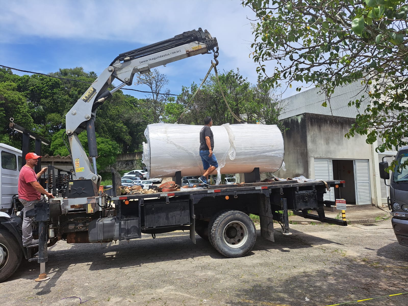 Dois homens trabalham no descarregamento de um grande cilindro industrial embrulhado em plástico, usando o guindaste acoplado a um caminhão. Um homem, de boné vermelho, opera o guindaste, enquanto o outro, de camiseta preta e bermuda jeans, está sobre a carroceria auxiliando no posicionamento da carga. Ao fundo, há árvores, carros estacionados e um prédio com paredes parcialmente encardidas e portas de metal.