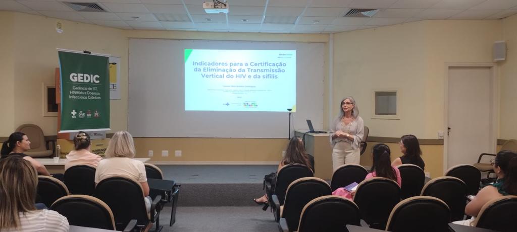 Mulher apresenta palestra em auditório para um pequeno grupo de pessoas. No telão, o slide exibe o título “Indicadores para a Certificação da Eliminação da Transmissão Vertical do HIV e da sífilis”. À esquerda, há um banner verde com o logotipo do GEDIC (Gerência de IST, HIV/Aids e Doenças Infecciosas Crônicas). O ambiente é claro, com cadeiras organizadas em fileiras e projetor ligado.