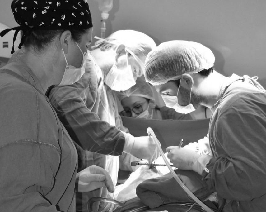 Fotografia em preto e branco mostrando equipe médica em um centro cirúrgico durante procedimento. Quatro profissionais, usando gorros, máscaras e aventais estéreis, trabalham concentrados sobre o paciente, manuseando instrumentos cirúrgicos. Ao fundo, há um suporte de soro pendurado.