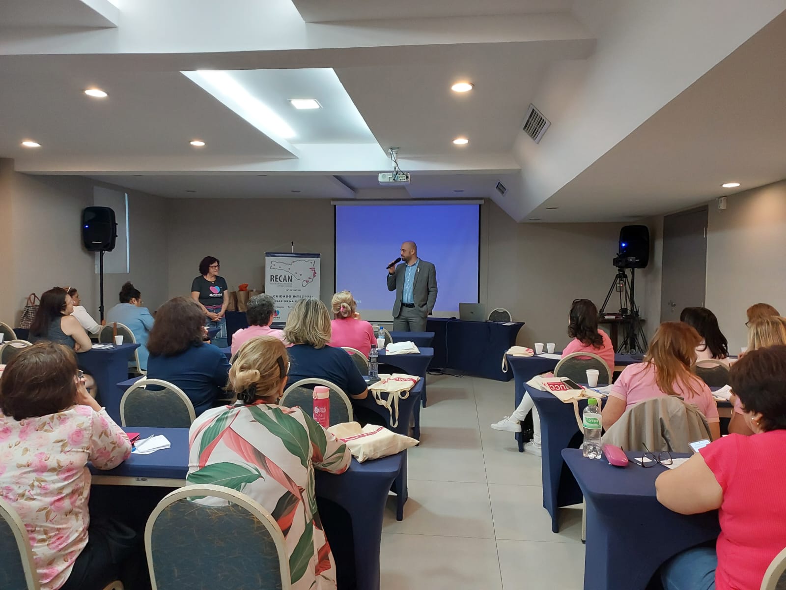 Fotografia de uma sala de conferência com diversas pessoas sentadas em mesas cobertas por toalhas azuis, voltadas para a frente. Um homem de terno cinza fala em pé, segurando um microfone, enquanto uma mulher ao lado observa. Ao fundo, há um telão azul projetado, um banner com o logotipo "RECAN" e equipamentos de som e filmagem.