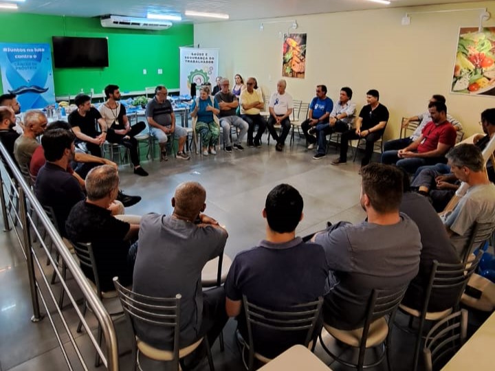 Grupo de cerca de vinte pessoas sentadas em círculo em uma sala, participando de uma roda de conversa. No fundo, há banners com mensagens sobre saúde do trabalhador e prevenção à Covid-19, além de mesas com garrafas de água e lanches. As paredes são decoradas com quadros de imagens de alimentos.