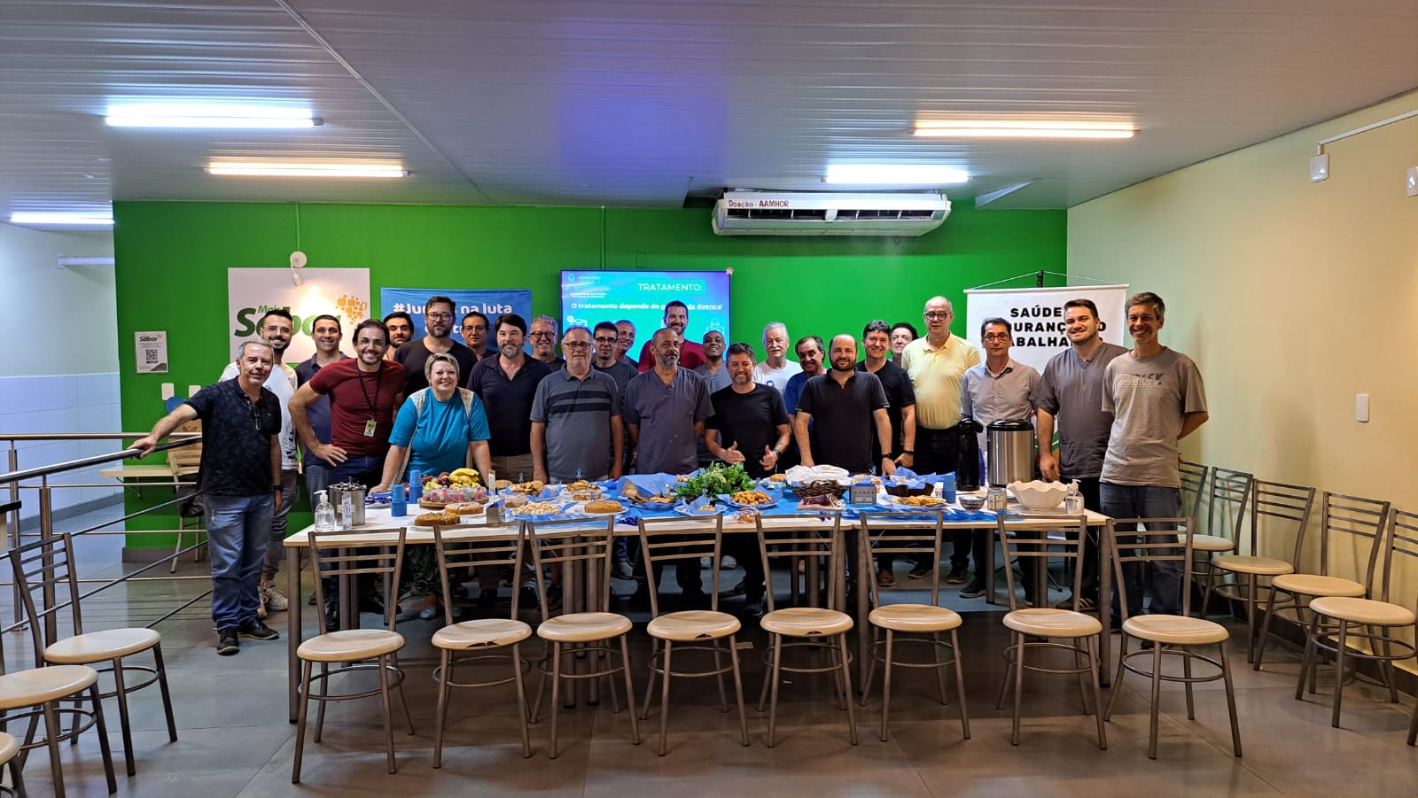 Grupo de cerca de vinte e cinco pessoas em pé, posando para foto em um ambiente interno com paredes verdes e amarelas. À frente, uma mesa grande coberta por toalha azul exibe café, pães, frutas e outros alimentos. Ao fundo, banners sobre saúde, segurança do trabalho e prevenção à Covid-19 estão expostos, junto a uma tela com a palavra “TRATAMENTO” em destaque.