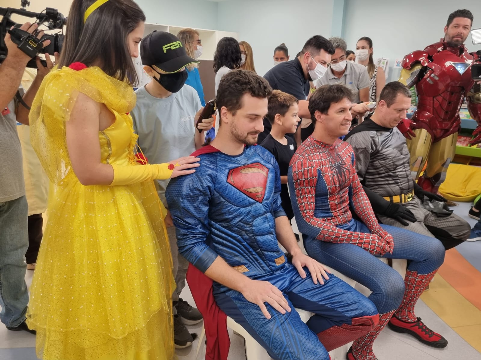 Grupo de pessoas fantasiadas de super-heróis e princesa participam de um evento interno. Três homens sentados, vestidos de Superman, Homem-Aranha e Batman, têm o cabelo cortado por voluntários, enquanto outras pessoas, incluindo um homem fantasiado de Homem de Ferro, acompanham e interagem. Há câmeras registrando a cena e algumas pessoas usam máscaras.