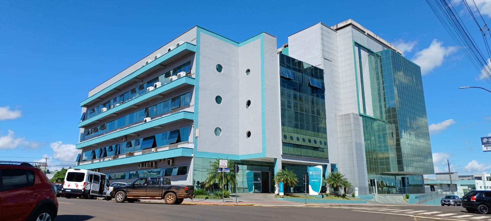 Fachada de um prédio moderno de vários andares, com detalhes em vidro espelhado verde e estrutura em tons de cinza e azul. Em frente, há veículos estacionados, incluindo ambulâncias e carros. O céu está azul com algumas nuvens, e há placas e vegetação próxima à entrada principal.