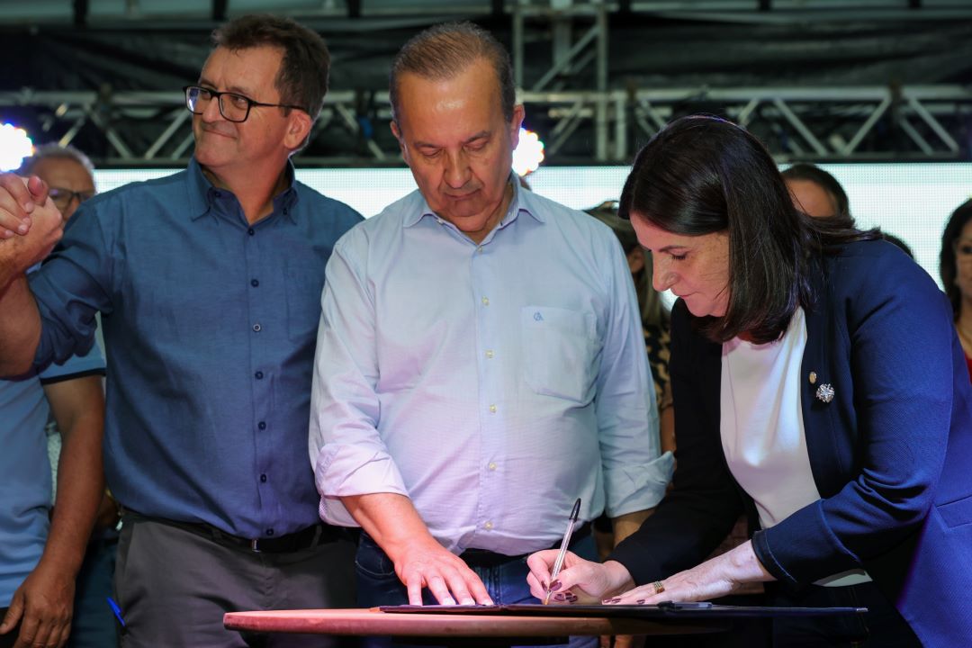 Mulher de blazer azul marinho e blusa branca assina um documento sobre uma mesa pequena, usando uma caneta. Ao seu lado, um homem de camisa azul clara observa atentamente, enquanto outro homem de óculos e camisa azul escura olha para o público, sorrindo. Ao fundo, outras pessoas acompanham a cena.