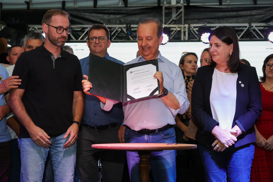 Grupo de pessoas em um evento oficial. No centro, um homem de camisa azul segura e exibe um documento aberto em uma pasta preta, enquanto sorri. Ao seu lado, um homem de óculos com camiseta preta e uma mulher de blazer azul marinho e blusa branca observam. Ao fundo, outras pessoas acompanham a cena.