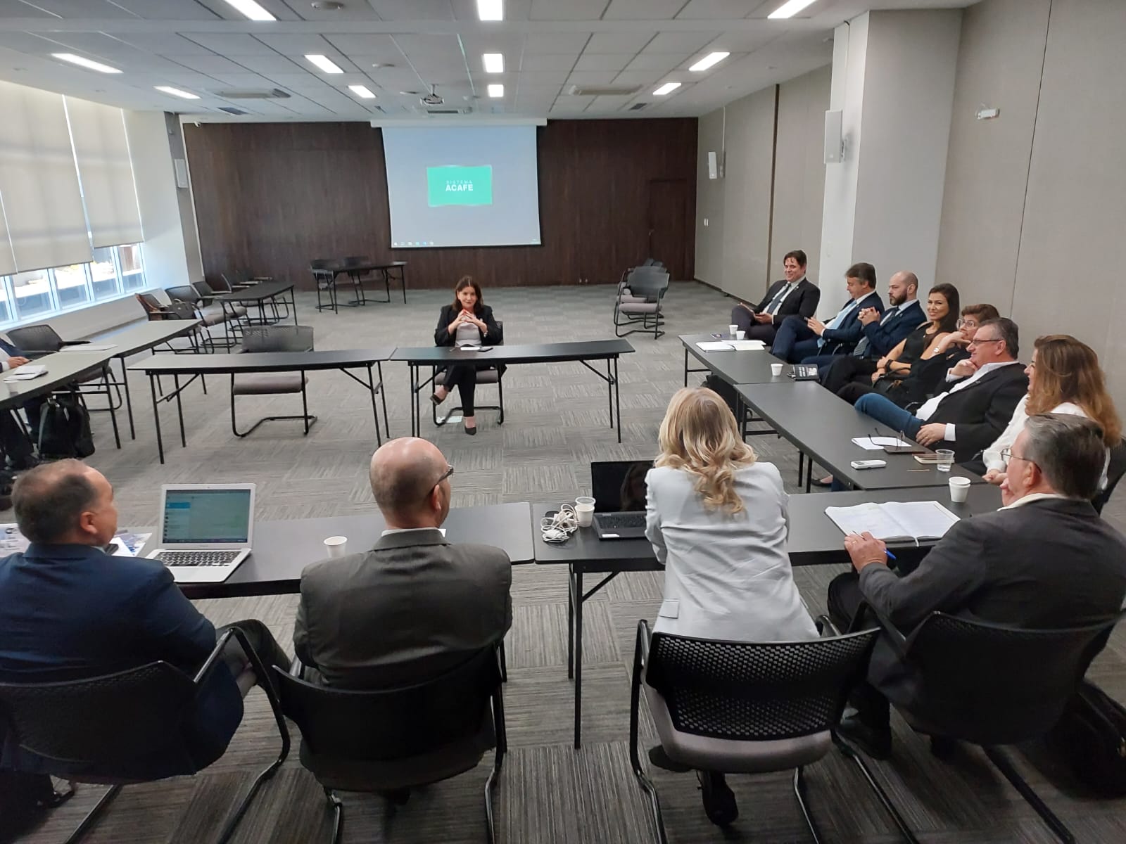 A imagem mostra uma reunião formal em uma sala ampla e bem iluminada, com mesas organizadas em formato de “U”. Ao centro, na parte frontal, está uma mulher sentada sozinha, conduzindo ou apresentando a discussão. Ao redor das mesas, cerca de dez participantes — homens e mulheres, a maioria em trajes formais — acompanham atentamente, alguns com notebooks, papéis e copos de café à frente.  No fundo da sala, há um projetor exibindo na tela a logomarca verde da ACAFE. O ambiente transmite um clima de reunião executiva ou encontro estratégico, com foco em diálogo e planejamento.