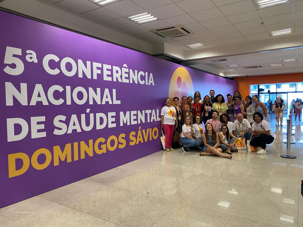 A imagem mostra um grupo de aproximadamente 25 pessoas posando para uma foto em frente a um grande painel roxo com letras brancas e amarelas que anunciam a "5ª Conferência Nacional de Saúde Mental – Domingos Sávio".  O grupo, formado por homens e mulheres de diferentes idades, está sorridente e aparenta estar participando do evento. Alguns usam camisetas brancas personalizadas, enquanto outros vestem roupas casuais ou sociais.  O ambiente é interno, com piso claro e brilhante, teto com iluminação embutida e entrada envidraçada ao fundo, permitindo a entrada de luz natural. O clima geral da foto é descontraído e celebrativo, típico de um registro de participação em congresso ou conferência.