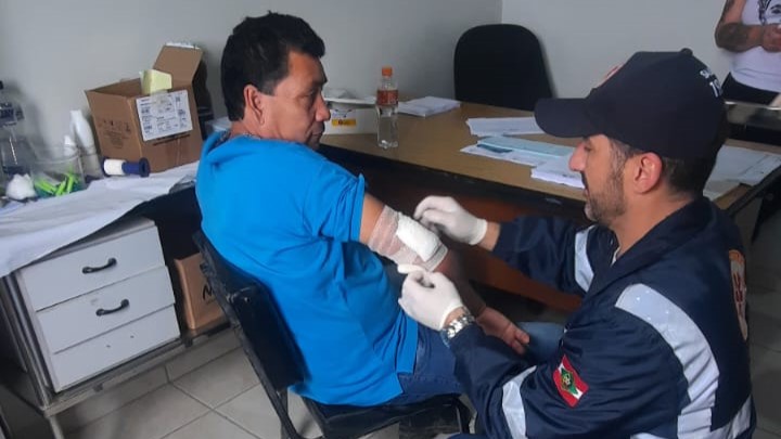 Profissional de saúde realiza curativo no braço de um homem sentado, durante atendimento de primeiros socorros em uma sala com mesa e materiais médicos.