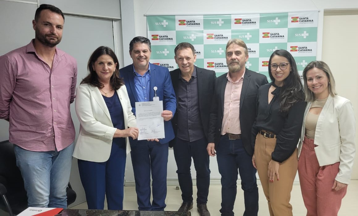 Sete pessoas posando para foto em frente a painel do SUS e Governo de Santa Catarina, enquanto duas delas seguram um documento oficial assinado.