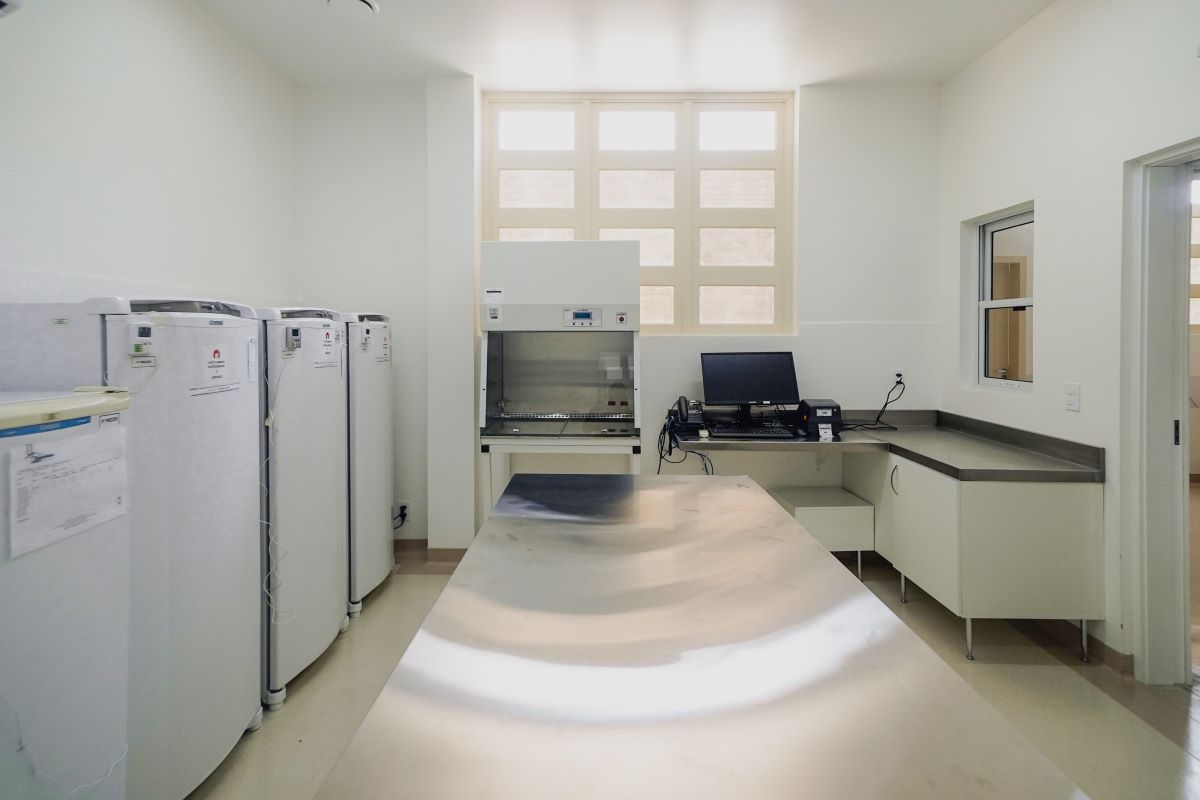 A imagem mostra um ambiente limpo e bem iluminado, provavelmente uma sala de laboratório ou de manipulação de alimentos/leite, equipada com superfícies e equipamentos adequados para trabalhos que exigem higiene e controle. À esquerda, há quatro refrigeradores verticais brancos, alinhados lado a lado. Ao fundo, no centro, encontra-se uma cabine de fluxo laminar, utilizada para manipulação asséptica, e à direita há uma bancada em “L” com um computador, telefone e impressora. No centro da sala, há uma mesa de aço inoxidável, refletindo a iluminação natural que entra por uma janela grande ao fundo. O espaço transmite sensação de organização, esterilidade e funcionalidade.