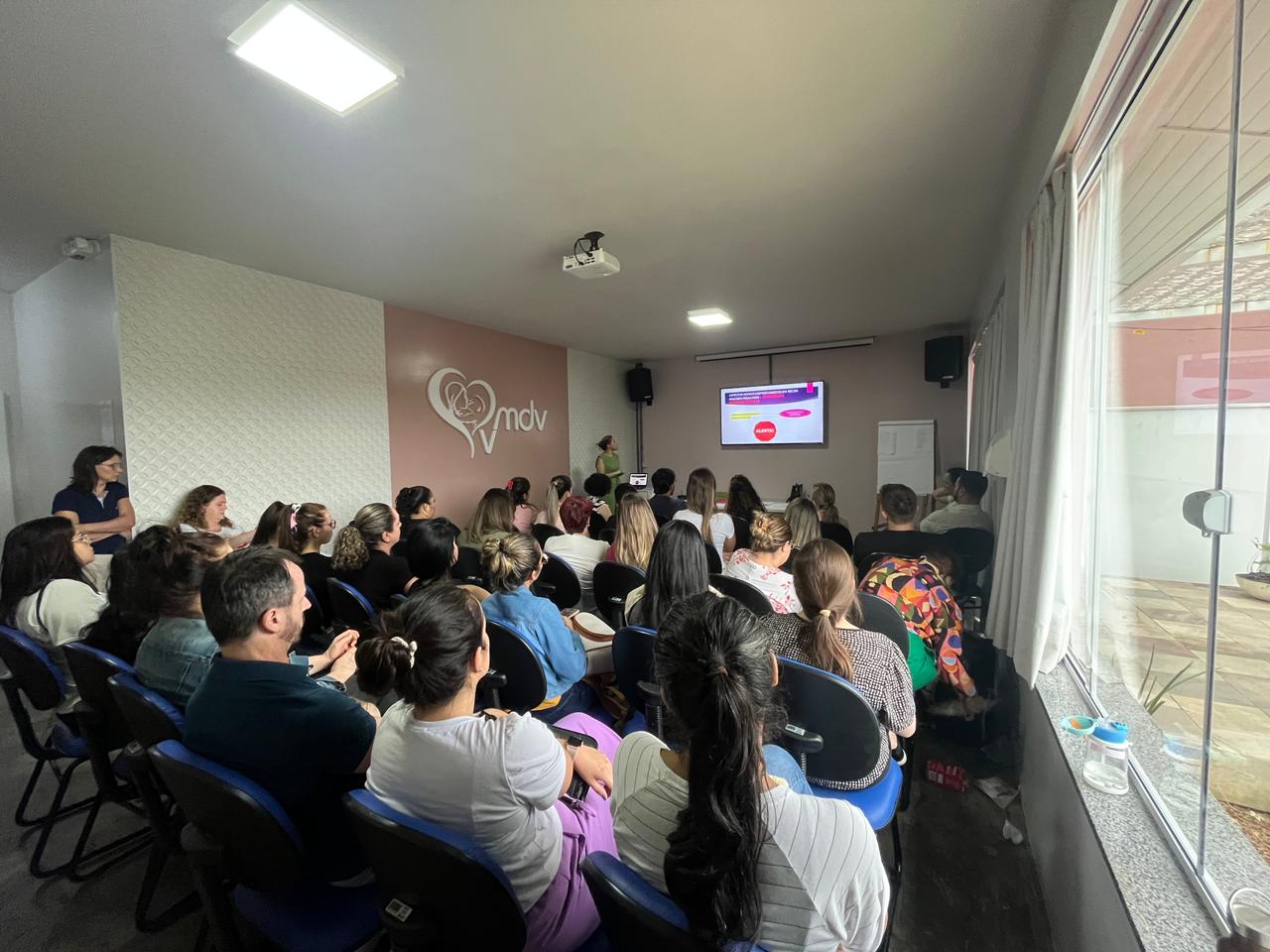 Sala de aula ou auditório pequeno com cerca de 30 pessoas sentadas em cadeiras azuis, assistindo a uma apresentação projetada em uma tela na parede frontal. A palestrante está de pé ao lado da tela, que exibe um slide colorido com textos e diagramas circulares. Na parede lateral há o logotipo do MDV, formado por um coração estilizado envolvendo o perfil de um bebê. À direita, uma grande janela deixa entrar luz natural.