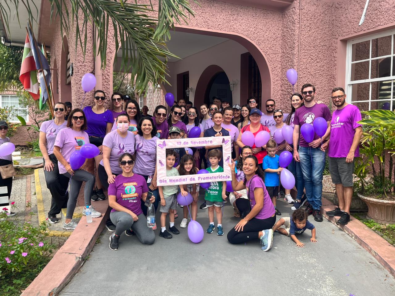 Grupo de homens, mulheres e crianças posando em frente a um prédio rosa, segurando balões roxos e usando camisetas predominantemente roxas em apoio à campanha do Dia Mundial da Prematuridade, celebrado em 17 de novembro. No centro, algumas crianças seguram uma moldura decorada com borboletas roxas e a frase 'Dia Mundial da Prematuridade'. Ao fundo, há bandeiras hasteadas e um arco de entrada para o edifício.